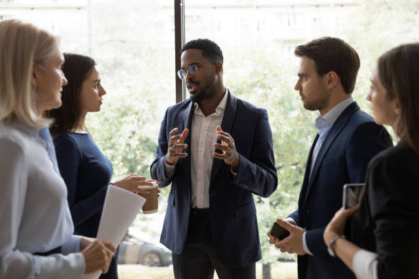 Confident African male leader standing telling diverse colleagues about new project, boss lead briefing for different age workgroup team employees in office. Sharing information and leadership concept